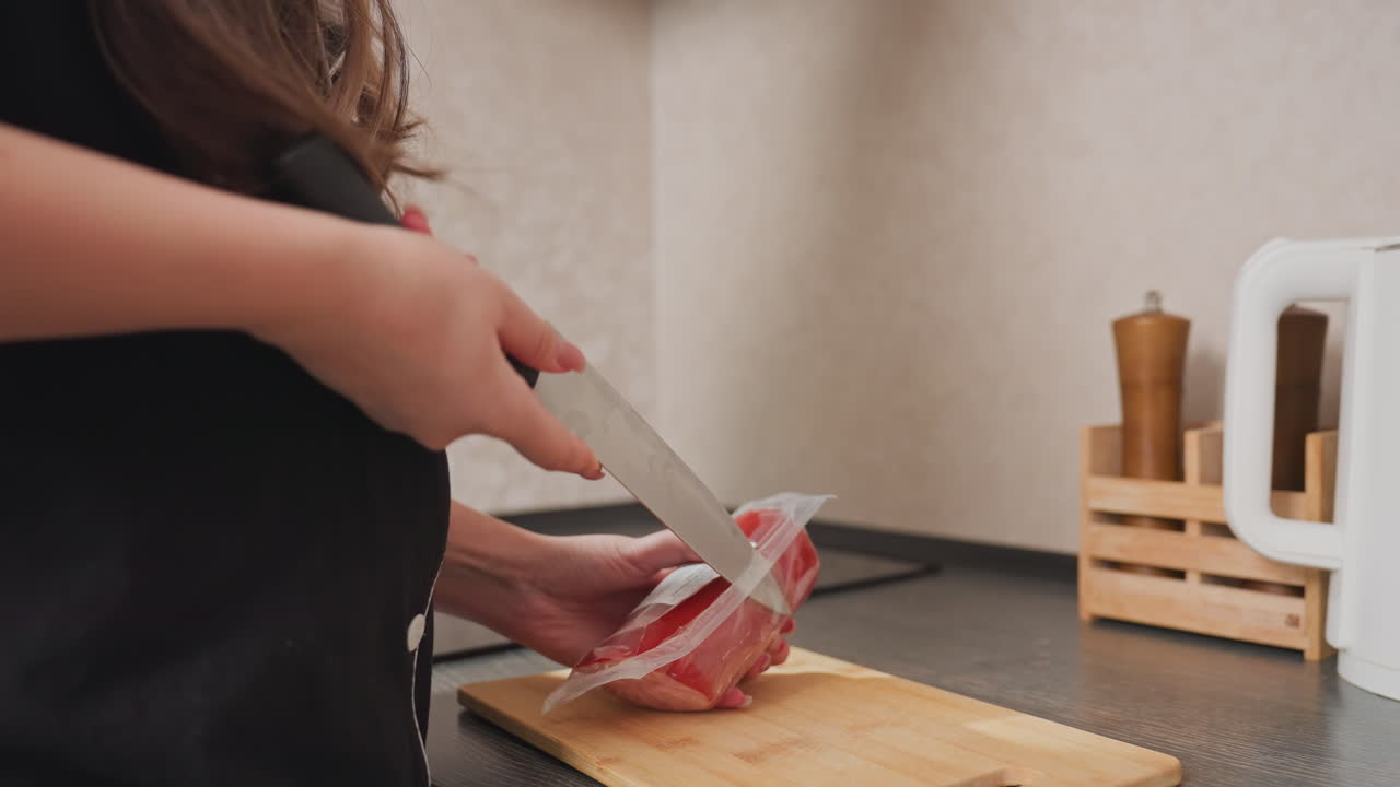 close up of chef using knife to slice open packaged hotdog placed on wooden cutting board on kitchen counter during meal preparation with spice containers and electric kettle in background