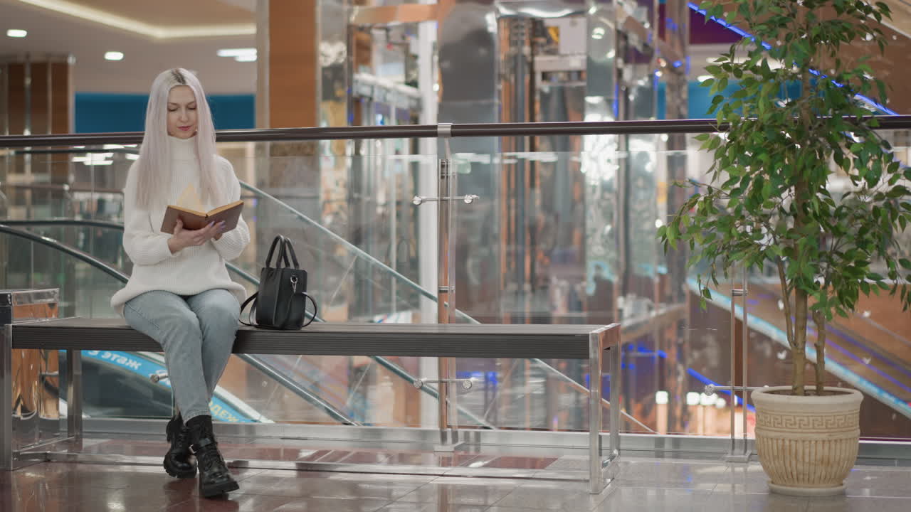 book lover walking mall walkway towards bench by decorative indoor tree, reaches into black handbag to retrieve hardcover book with elegant hands, prepares to sit and read amid retail atmosphere
