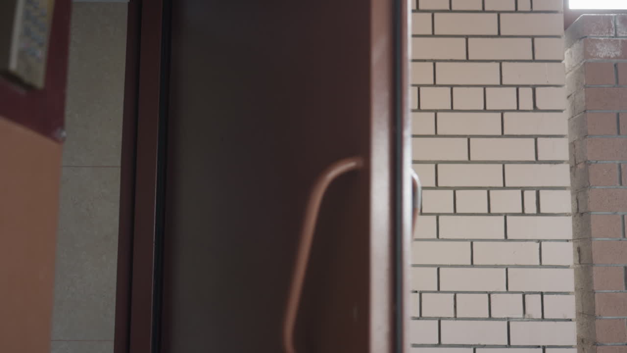 Стильный профессионал открывает дверь безопасности и выходит наружу, минуя панель с клавиатурой, видны коричневая металлическая рама и кирпичный коридор, размытое движение на солнцезащитных очках и шарфе, яркий дневной свет