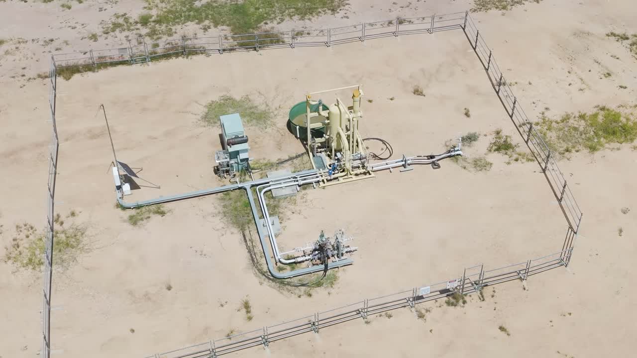 Aerial perspective of a fenced gas extraction site with visible machinery and pipelines on a dirt field.
