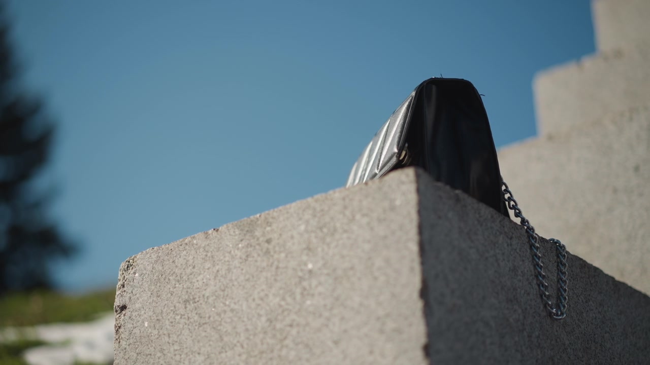 Close-up van een zwarte leren handtas met kettingriem, rustend op een betonnen blok onder fel zonlicht. De achtergrond toont een helderblauwe lucht en een ver groen landschap, wat de textuur van de tas benadrukt