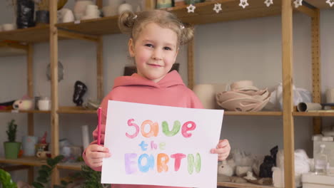 blonde little girl in craft workshop holding a cardboard sign with save the earth phrase