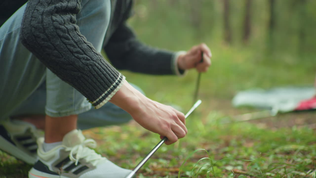 Close up of lady handing tent poles to man in canvas squatting in forest as he joins them together for camping setup with soft natural light and blur background