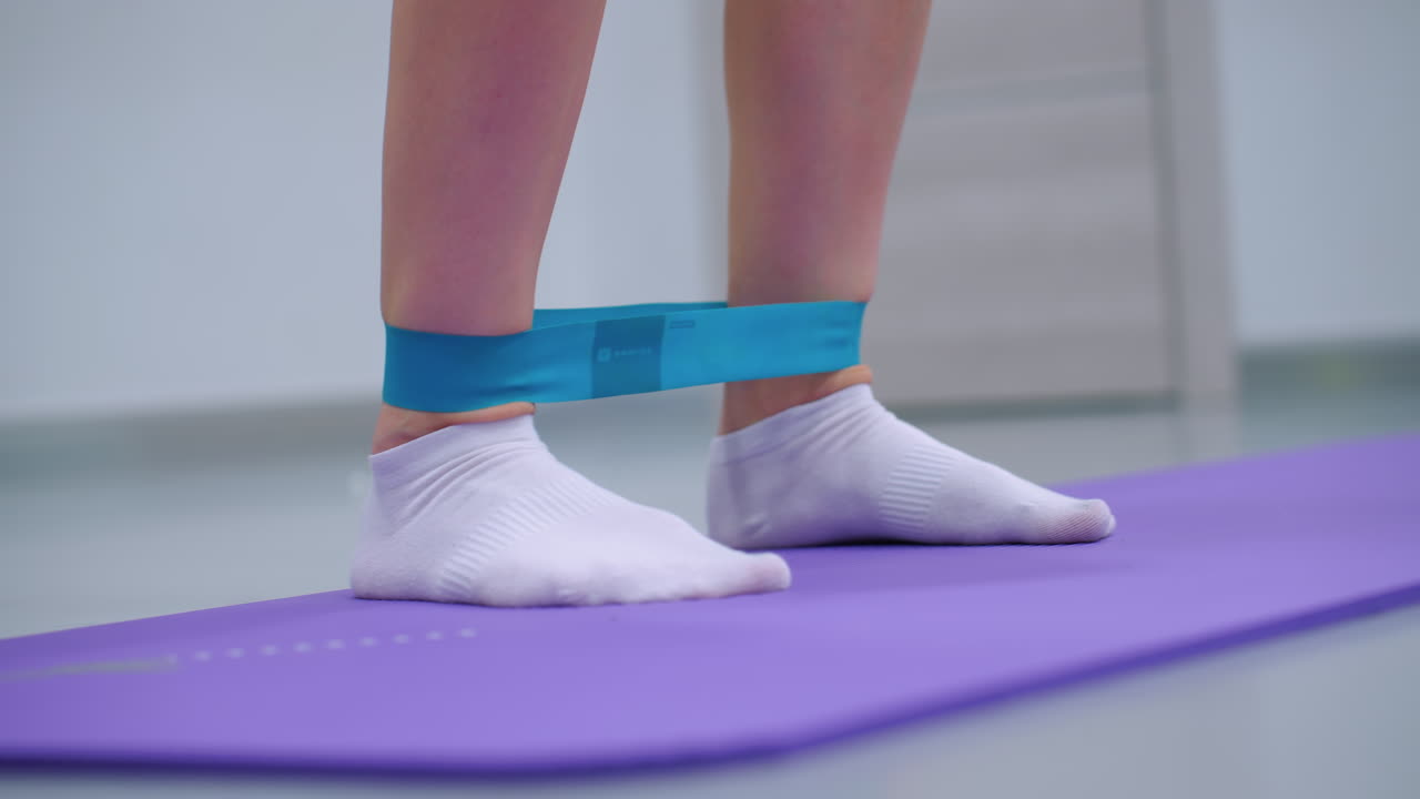 vista de ángulo inferior de primer plano de una persona con calcetines blancos usando una banda de resistencia para estiramientos laterales de las piernas, la alfombra de ejercicio púrpura y la pelota de gimnasia verde en el fondo