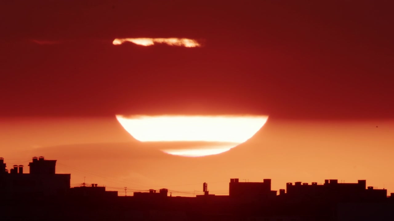 Wide shot of the sun slowly lowering behind a dense band of dark clouds, with a glowing horizon line beneath