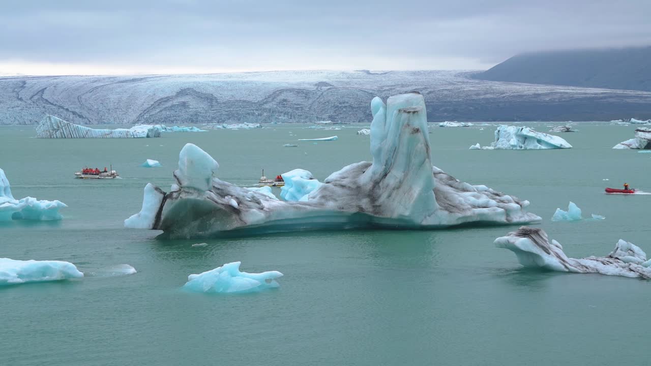 상대적으로 작은 보트가 요쿨살론의 청록색 빙하 호수 물에서 거대한 빙산을 지나갑니다.