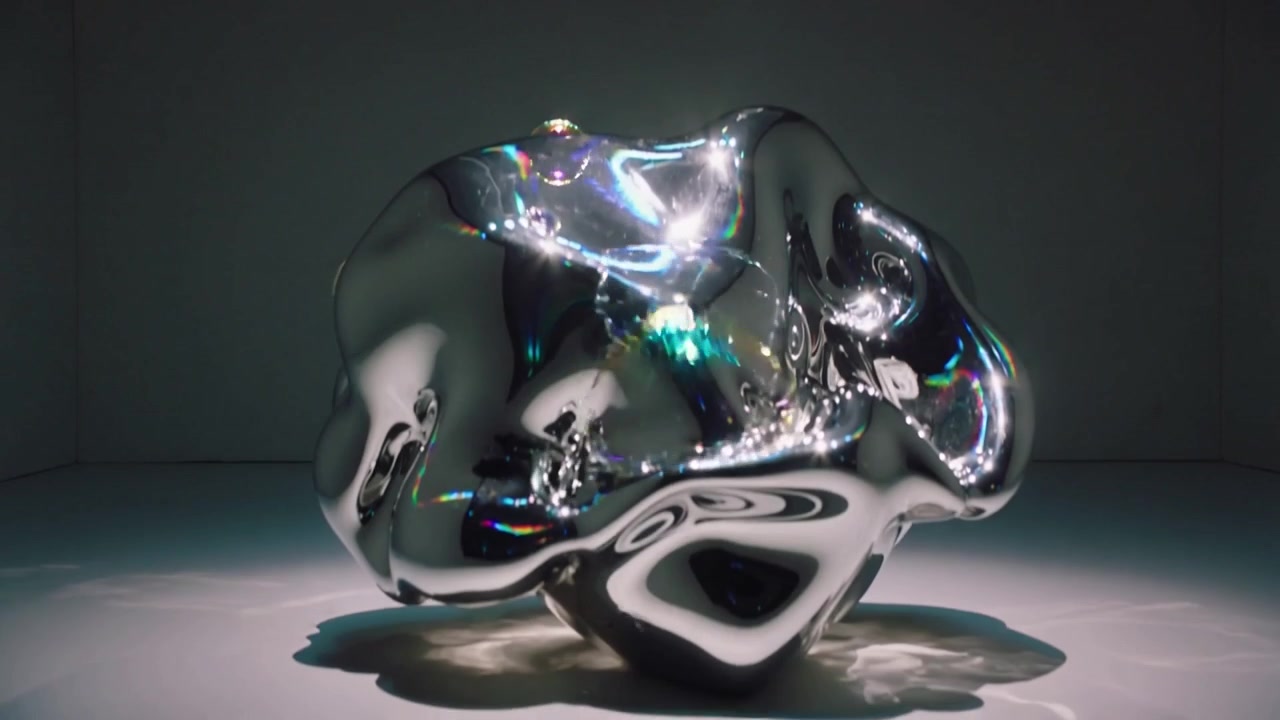 Tre bolle in caduta verso una scultura cromata in studio, una che scoppia, lasciando una pellicola arcobaleno.