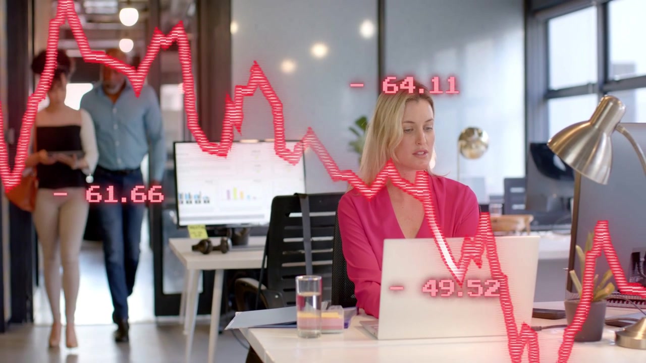 Grafico azionario rosso che si muove su un laptop, con una donna che monitora il mercato, digita e reagisce mentre il team passa.