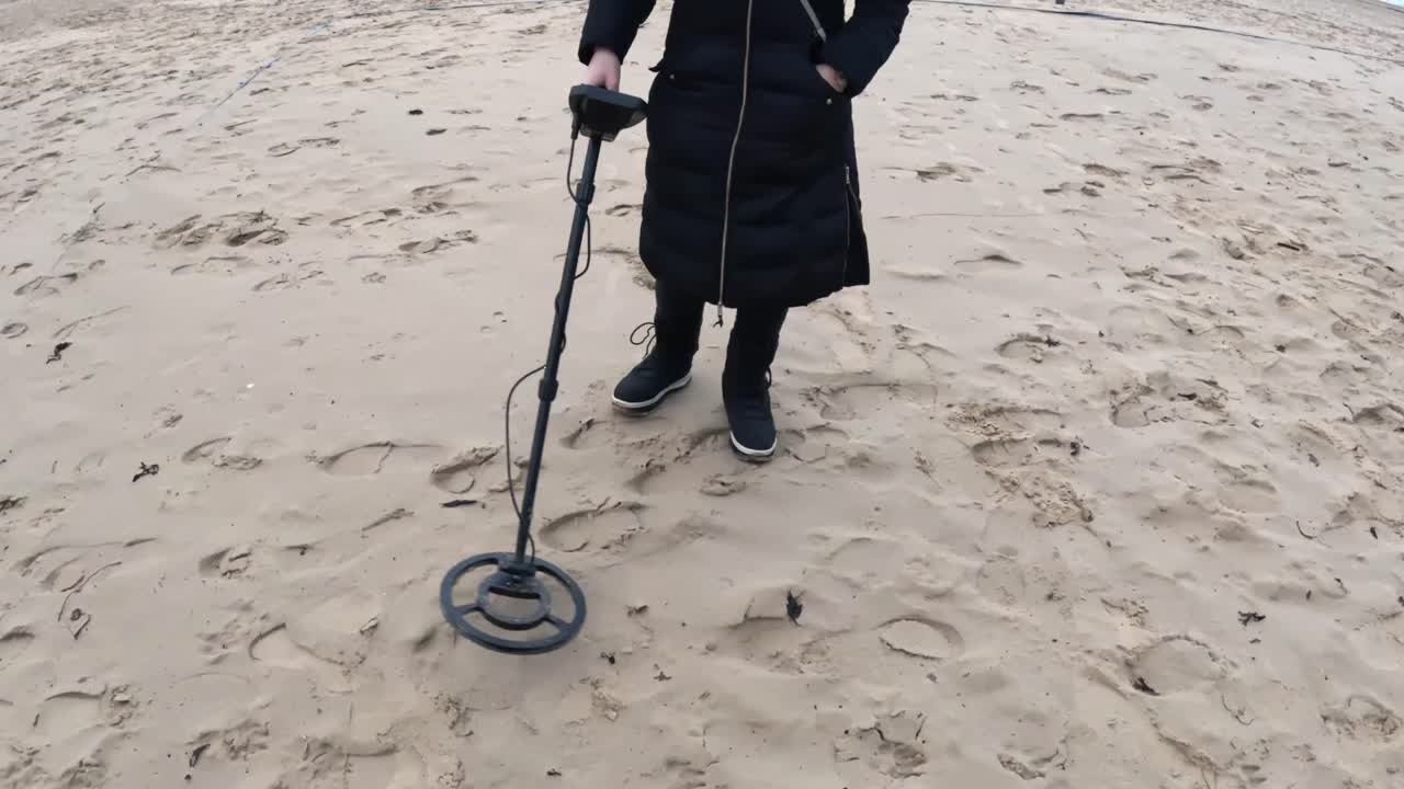 Persona che utilizza un metal detector sulla sabbia soffice della spiaggia vicino a un nastro blu strappato che delimita l'area, che si muove lentamente mentre scansiona le impronte alla ricerca di oggetti metallici smarriti