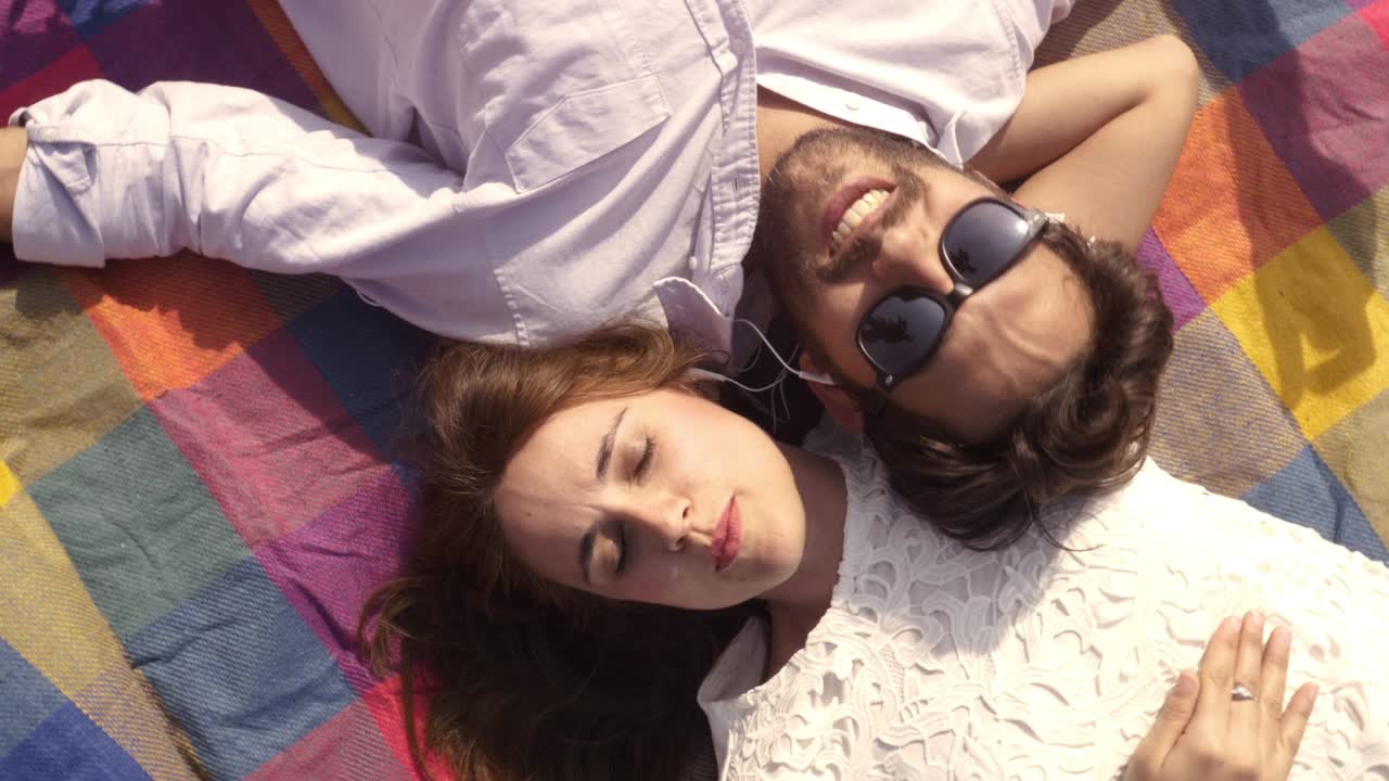 행복하고 사랑스러운 젊은 부부는 공원에서 다채로운 담요에 누워 스마트폰에서 음악을 듣고 헤드폰으로 기타 선글라스와 함께 로맨틱합니다. 아름다운 매력적인 소녀  뷰 회전 카메라 느린 동작