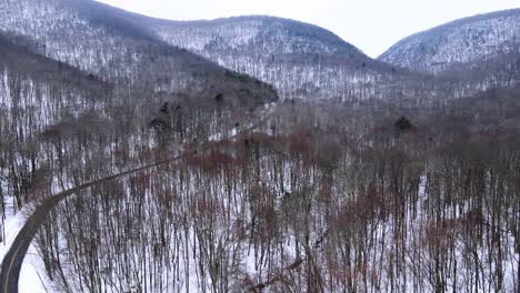 Flying-over-a-snowy-mountain-valley-with-forests-below-and-a-remote-mountain-highway,-and-snow-covered-mountains