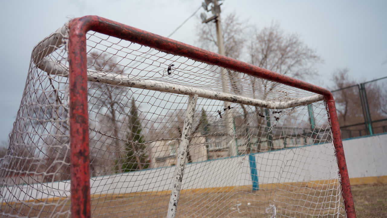 soccer ball hitting net midair. fast moving ball slams into red goalpost net on outdoor rink, blurred background with bare trees and fence, energy and motion captured, splintered paint and gritty