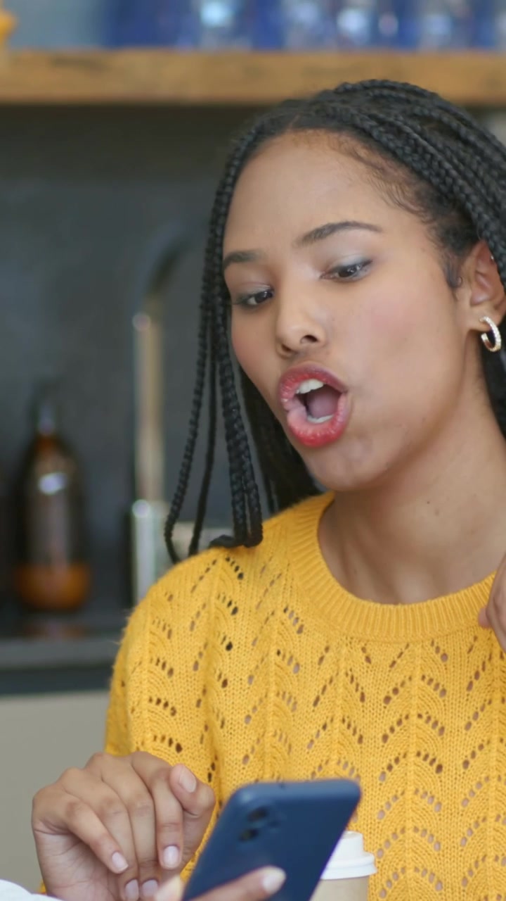 Vertical video: Woman in yellow top taking handed phone checking it with cup at counter, copy space