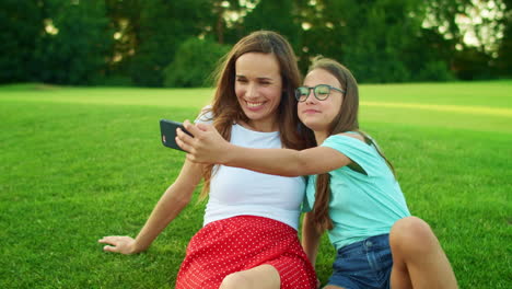 Girl-and-woman-talking-selfie-on-cellphone.-Daughter-and-mother-sitting-on-grass