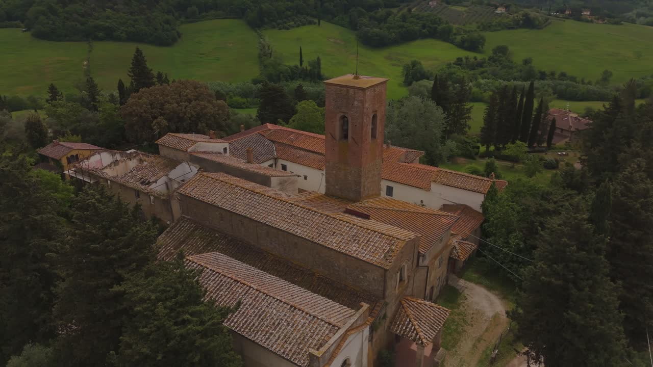 Drone shot of a medieval church complex with a tall brick bell tower, nestled in the green hills and olive groves of Tuscany. Peaceful rural setting with rustic charm and traditional architecture.
