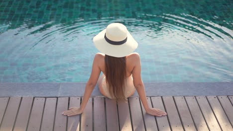 Una-Mujer-Joven-Con-Un-Sombrero-Para-El-Sol-Y-Un-Traje-De-Baño-Se-Sienta-De-Espaldas-A-La-Cámara-En-El-Borde-De-La-Terraza-De-Una-Piscina-Se-Inclina-Hacia-Atrás-Sobre-Sus-Brazos