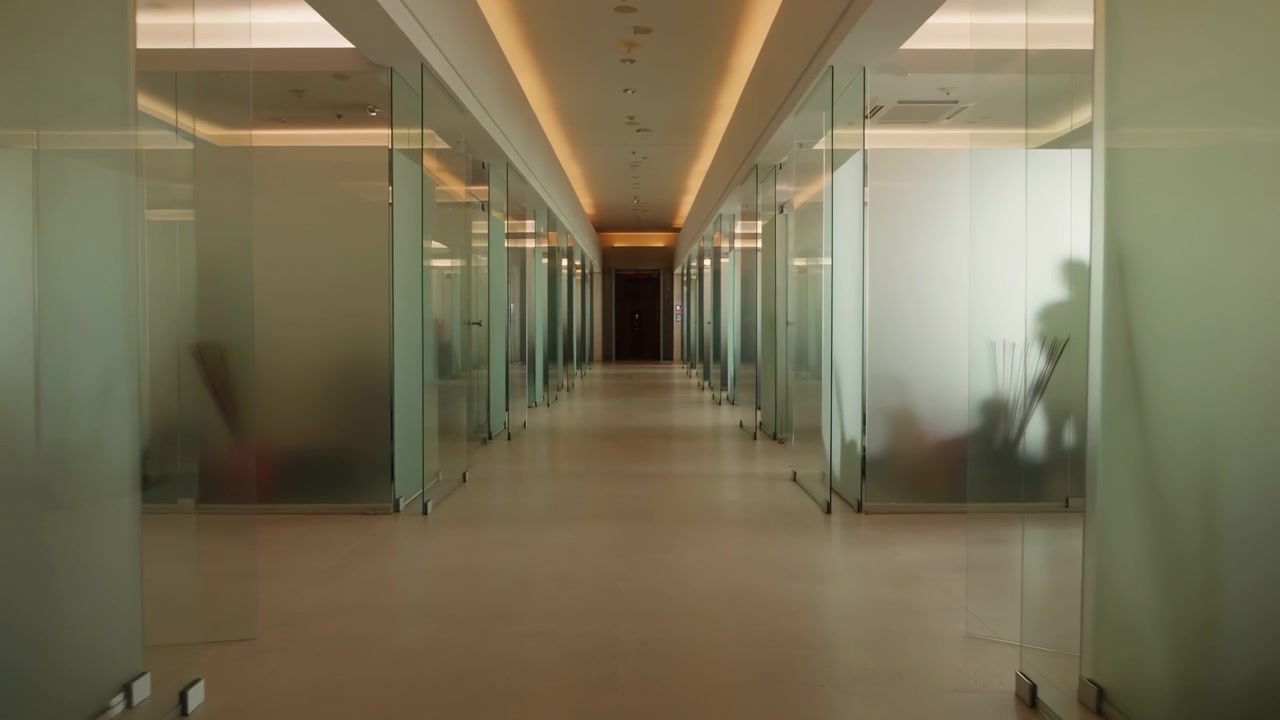 A câmera avança constantemente por um corredor simétrico enquanto paredes transparentes de salas passam em ambos os lados, com reflexos suaves, iluminação embutida quente, interiores discretos e uma porta distante à frente