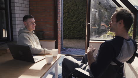 young man talking to his disabled friend in wheelchair during a sunny day at home