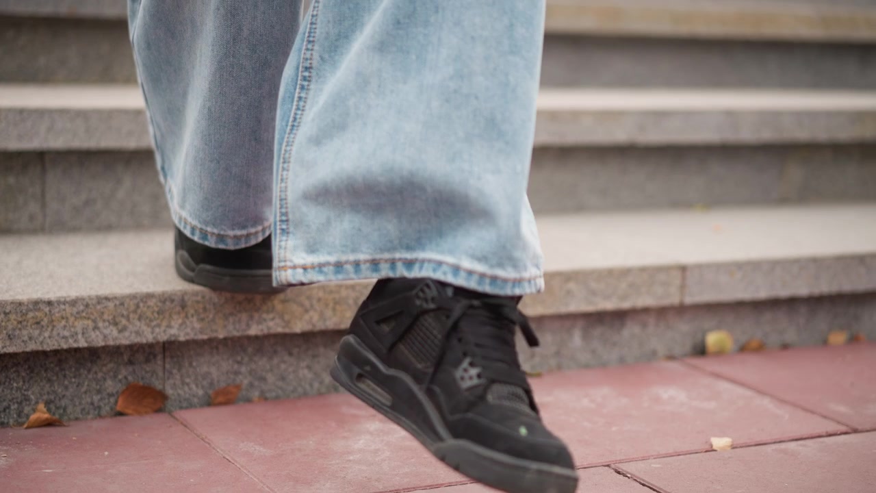 Primo piano dei piedi di una persona con scarpe da ginnastica nere e jeans azzurri che scende gradini di pietra con foglie marroni sparse a terra. Il fuoco rimane costantemente sulla parte inferiore delle gambe, sulle scarpe e sul gradino