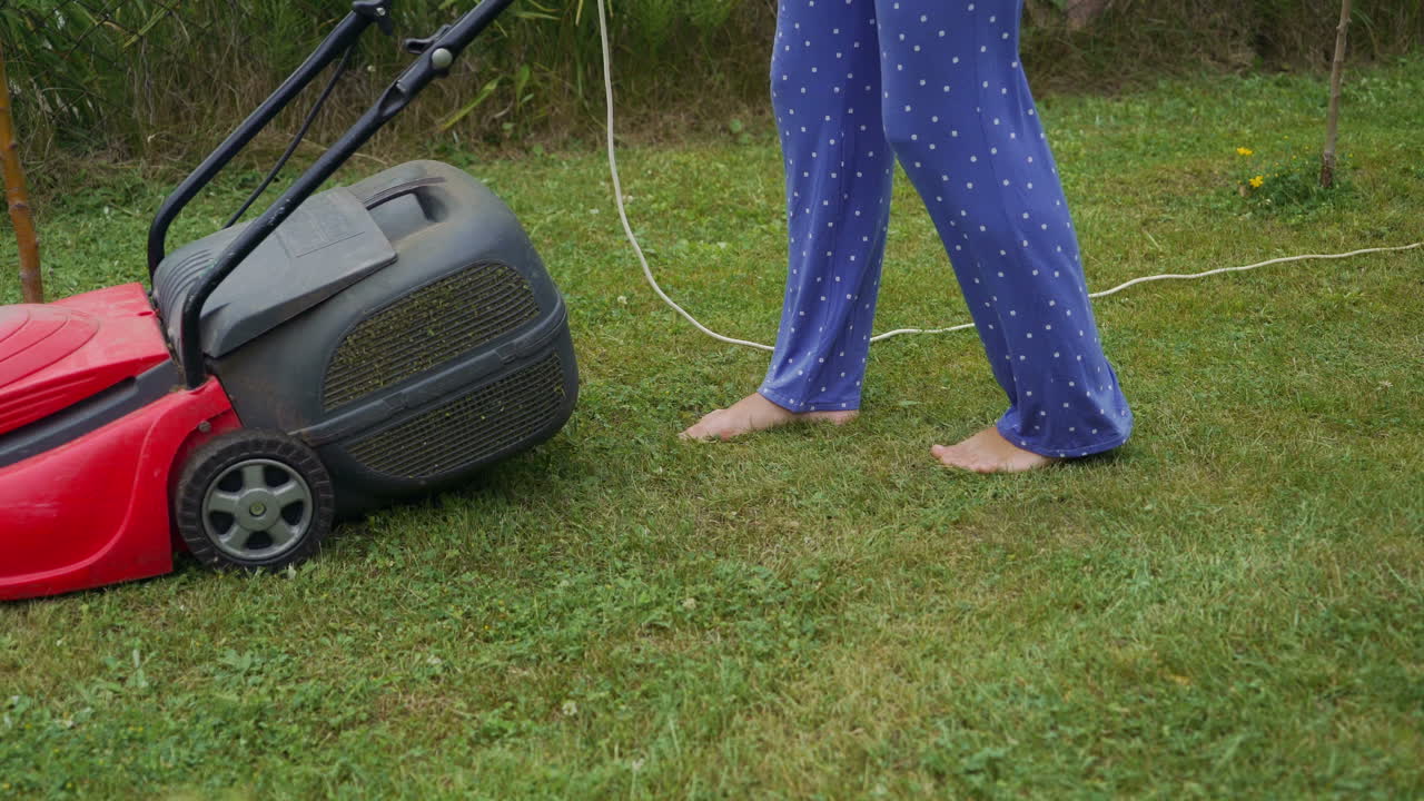 Barefoot young girl mowing grass with a lawn mower in garden. Bare female feet walking behind lawn mower in back yard