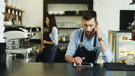 Junger-Hübscher-Kellner,-Der-Am-Telefon-Spricht-Und-Auf-Dem-Tablet-gerät-An-Der-Bar-Scrollt,-Hübsche-Kellnerin,-Die-Dahinter-Kaffee-Kocht