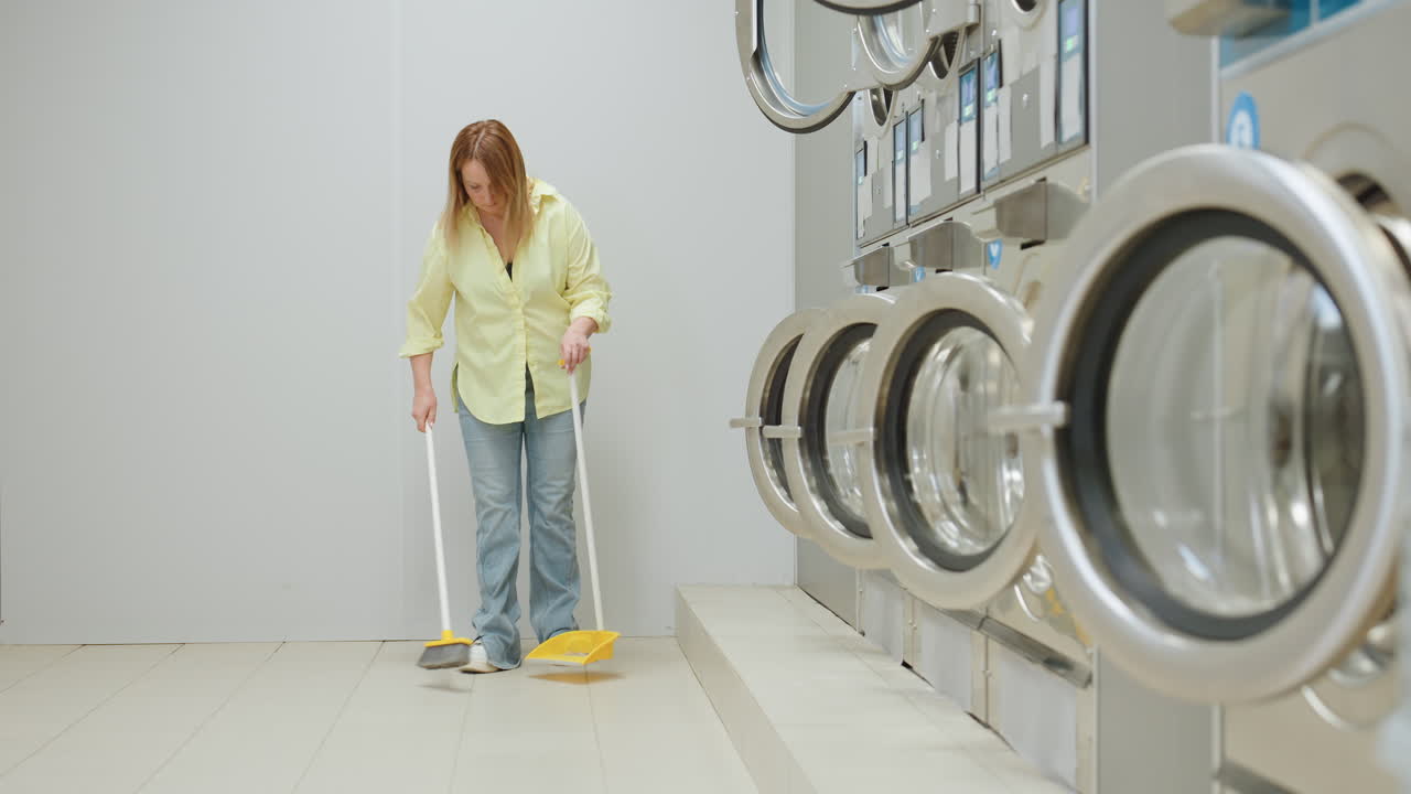 Zoom out shows woman near industrial washing machines in laundromat, yellow shirt and jeans with white sneakers sweeping tiled floor using broom and dust pan, routine cleaning in bright room