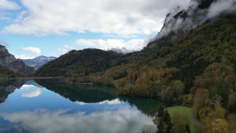 Volando-A-Lo-Largo-Del-Lago-Con-Agua-Clara-Situada-Entre-Un-Valle-Rodeado-De-Altos-Picos-Montañosos-Cielo-Nublado