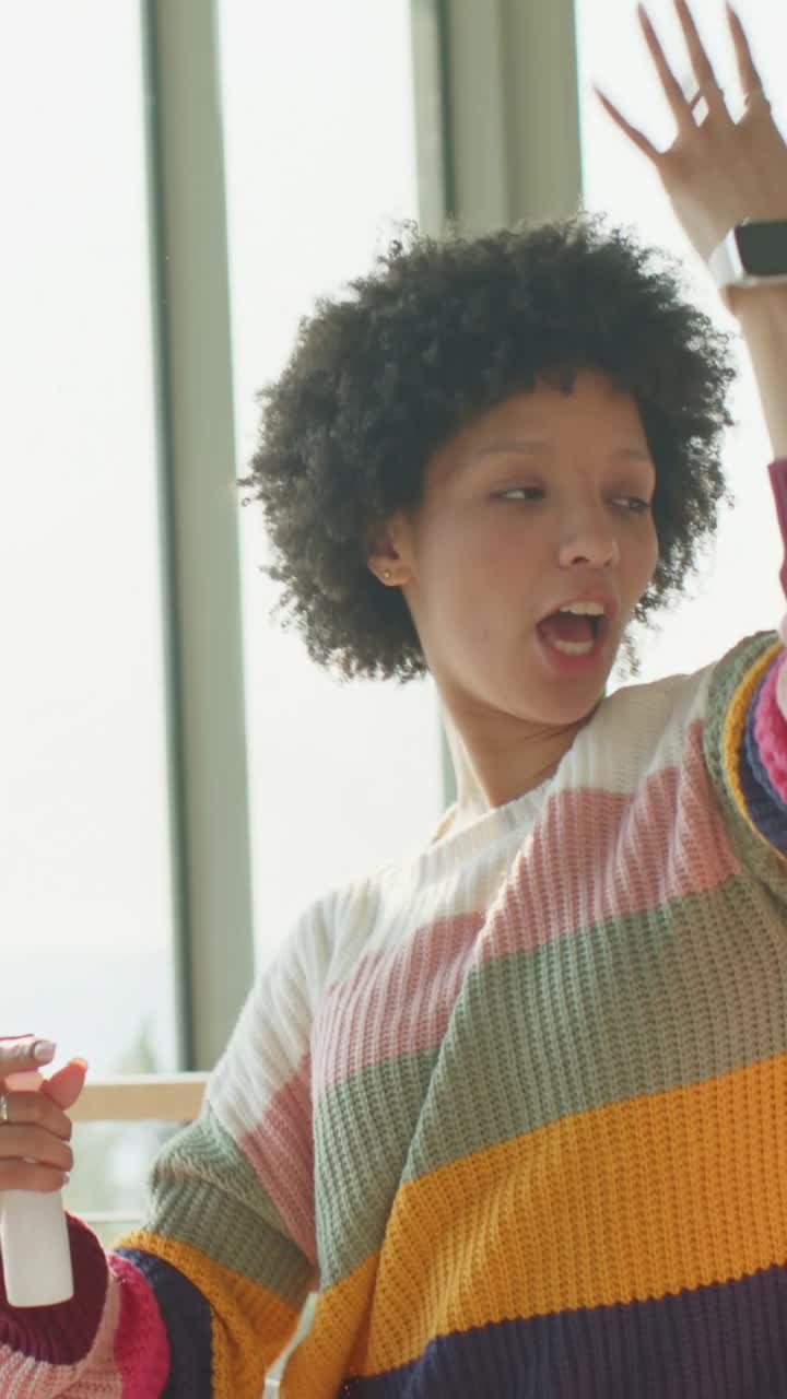 Vertical video: Raising arm, woman in striped knit dancing at home, showing pink bottle, singing
