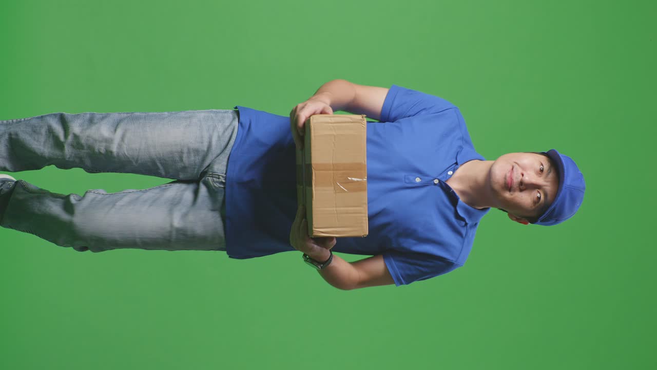 푸른 유니폼을 입은 미소 짓는 아시아 남성 택배기사가 스튜디오에서 녹색 스크린 배경에 상자를 배달하는 동안 걸어다니고 둘러보고 있습니다.