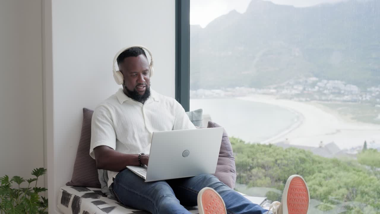 Adult man is typing on laptop at window bench, pausing, looking at bay, speaking on call