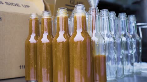 Filling-up-Cayenne-chilli-sauce-into-glass-bottles-with-metal-funnel-and-boxes-background