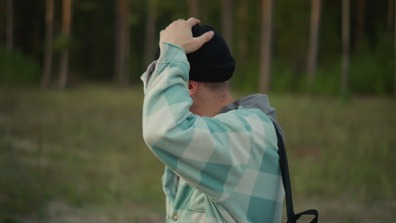 White hiker in forest clearing at dusk, facing camera briefly, adjusting beanie, scanning treeline, relaxed expression, layered flannel jacket, backpack straps visible, soft evening light, candid