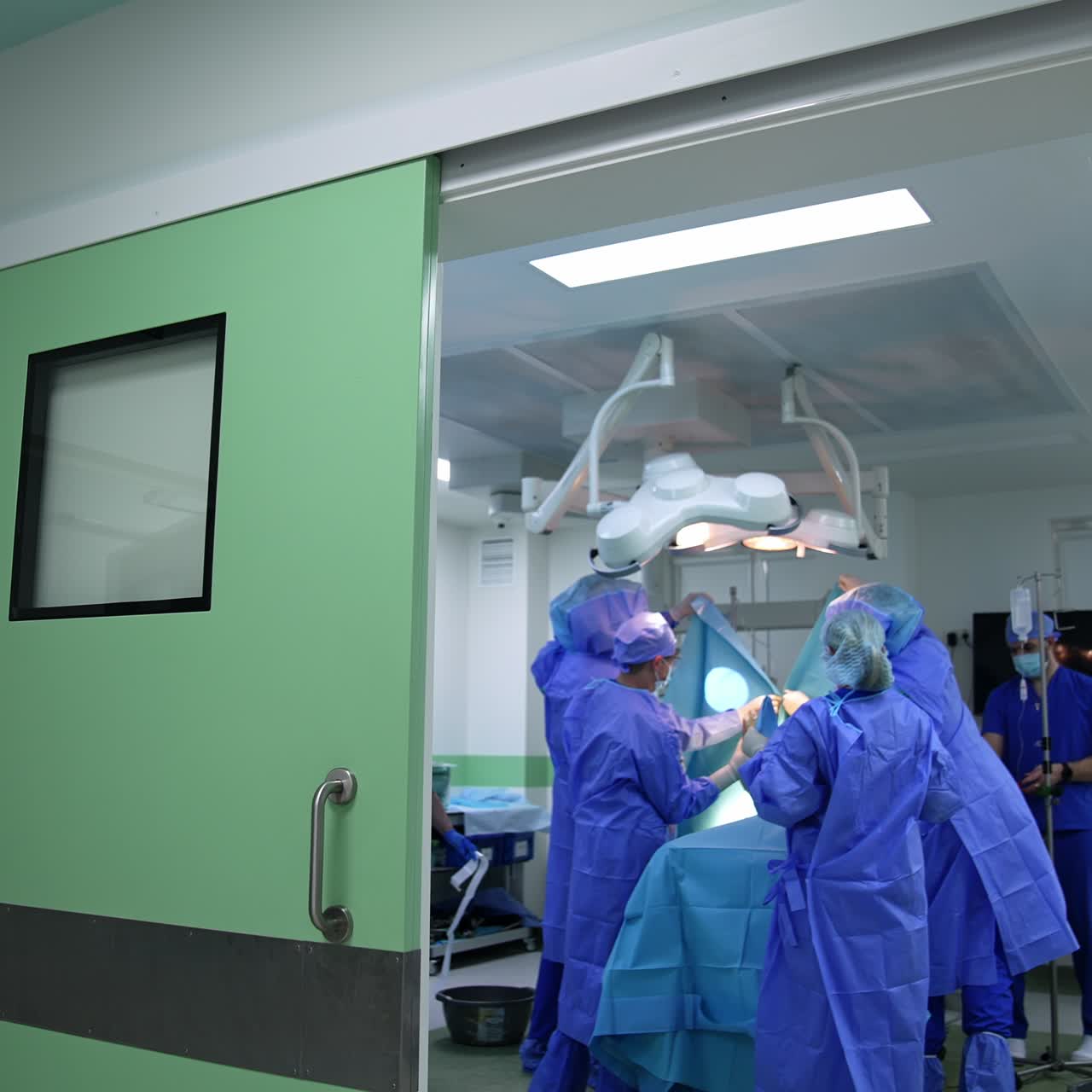 Large medical team of doctors working in the modern surgery room. Surgeons are covering the place of surgery with a sheet. Footage at the doors to an operational theatre