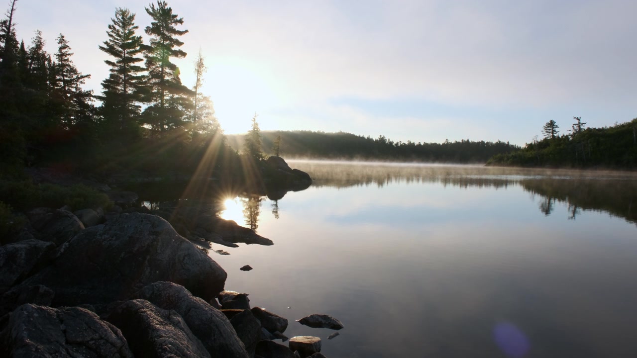 La luce del sole mattutina filtra tra i pini lungo una riva rocciosa mentre la nebbia si alza da un sereno lago selvaggio nelle Boundary Waters