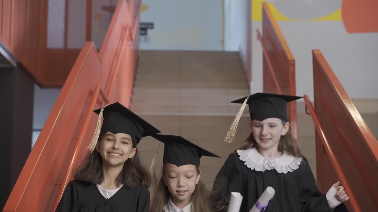 три счастливые студентки дошкольного учреждения в шапочках и платьях бегут по лестнице и держат диплом на выпускной церемонии