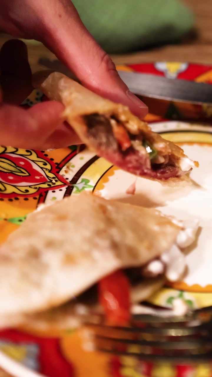 Una mano levanta una porción de pizza mexicana de un plato vibrante y estampado, mostrando ingredientes frescos en un ambiente cálido.