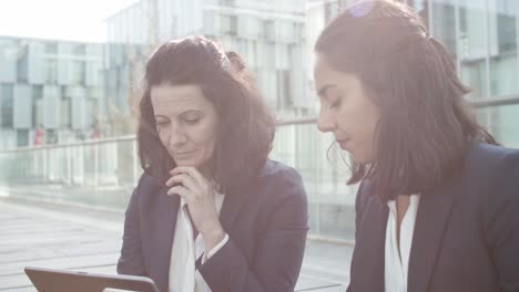 Serious-Businesswomen-Using-Laptops-For-Work-Together-While-Sitting-Near-Office-Building-Outside,-Discussing-Content-And-Pointing-At-Displays