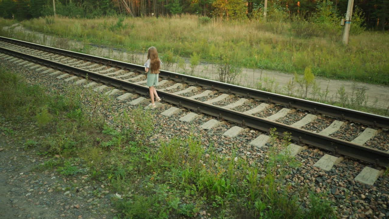 Lonely kid walking carefully along railway sleeper in quiet countryside, dressed in denim dress and striped shirt, surrounded by wild grass, gravel stones, and dirt road