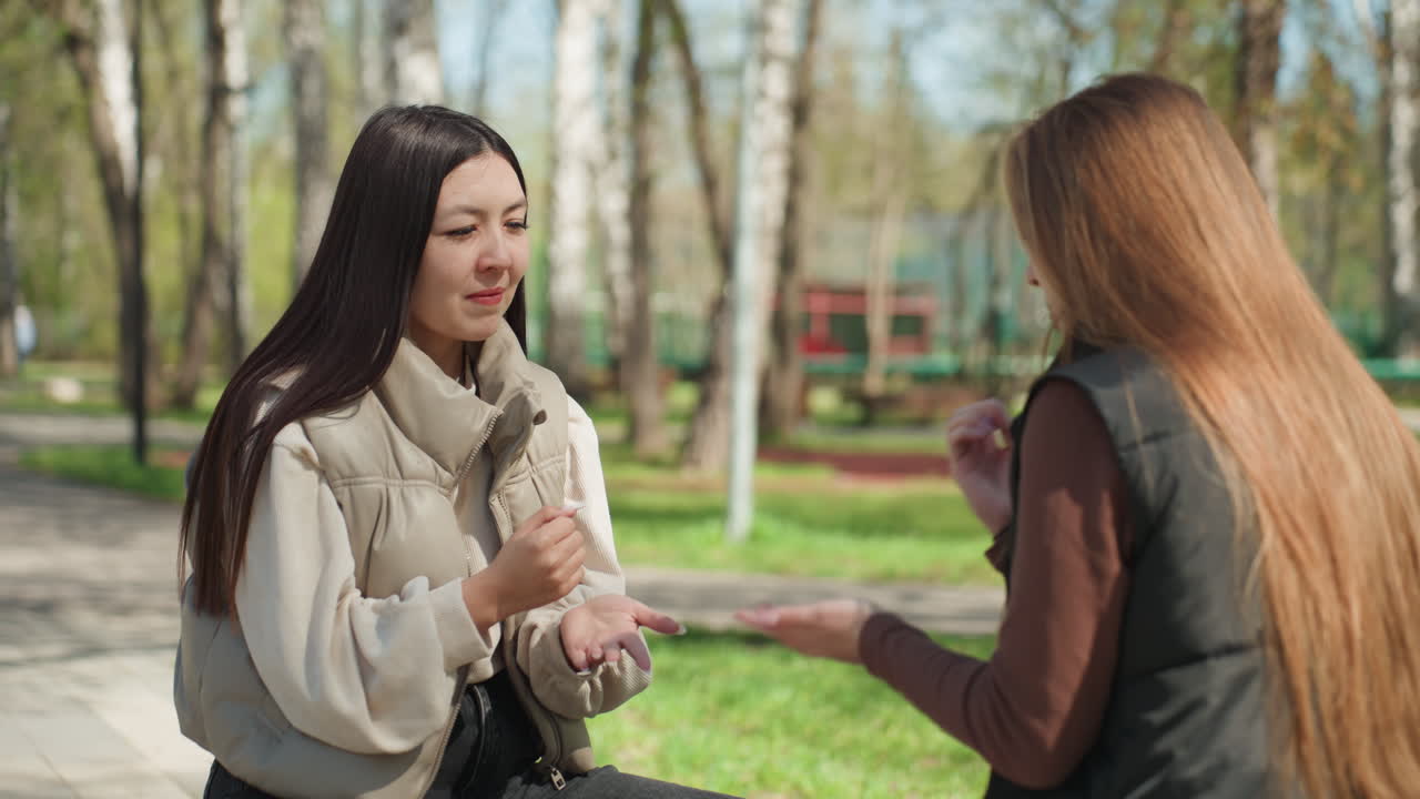 Người phụ nữ châu Á và người bạn da trắng trao đổi vòng tay trên ghế đá công viên, cây cối mùa xuân và ánh nắng mặt trời, áo khoác thường ngày, trò chuyện thân mật, biểu cảm tươi cười và đôi tay nhẹ nhàng trao món quà nhỏ, tự nhiên