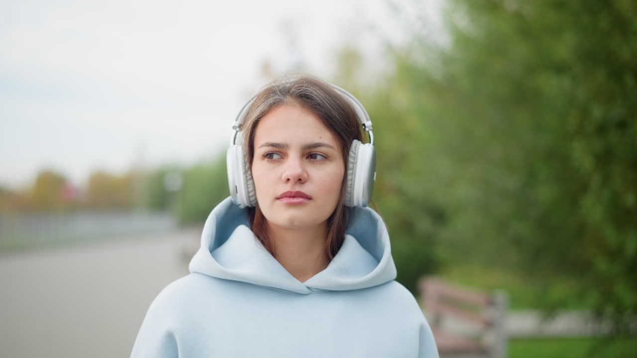 Close-up view of young woman strolling in garden wearing headphones, with blurred trees and bench in background. Perfect for videos about music enjoyment, relaxation, or nature walks