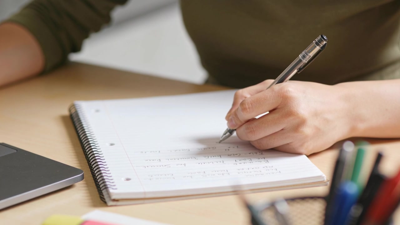 Pen touching paper, right hand writing notes in spiral-bound notebook at desk, olive sleeve