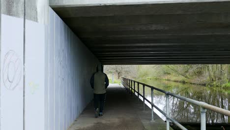 the back of an anonymous man walking under an overhead bridge, the footpath running parallel to the bank of the thetford little river in norfolk, england