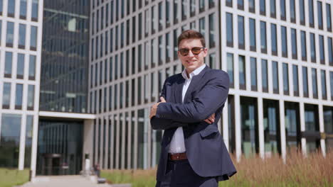 joven hombre de negocios en traje y gafas de sol posa frente al rascacielos