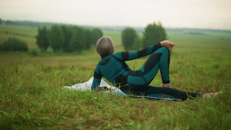 back view of a woman lying on one side with her hand on her leg, looking focused and relaxed in a grassy field, the trees and greenery in the background are slightly blurred