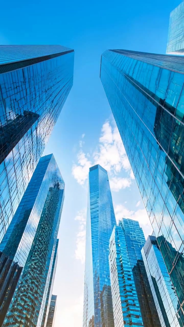 Vista de baixo ângulo entre altas torres de escritórios de vidro em uma cidade moderna, com fachadas reflexivas emoldurando um céu azul, nuvens dispersas e luz solar quente.