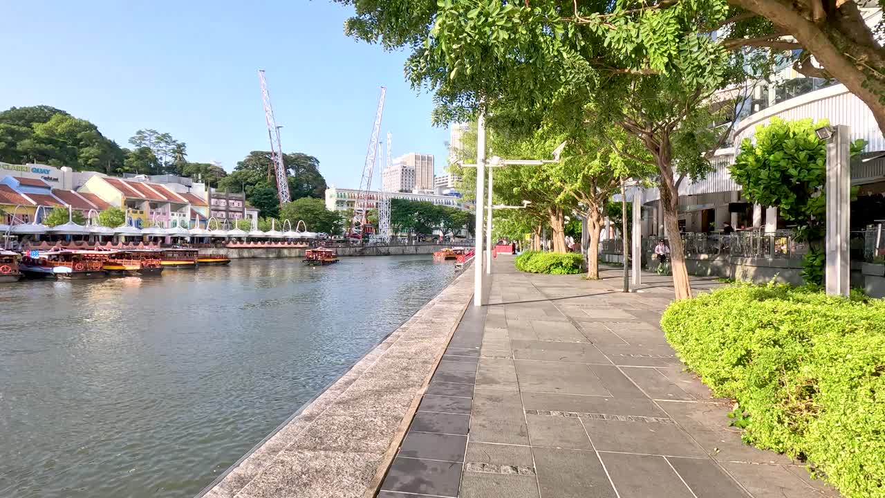 Daytime camera movement along Clarke Quay riverfront, colorful buildings, boats, lush greenery, clear sky