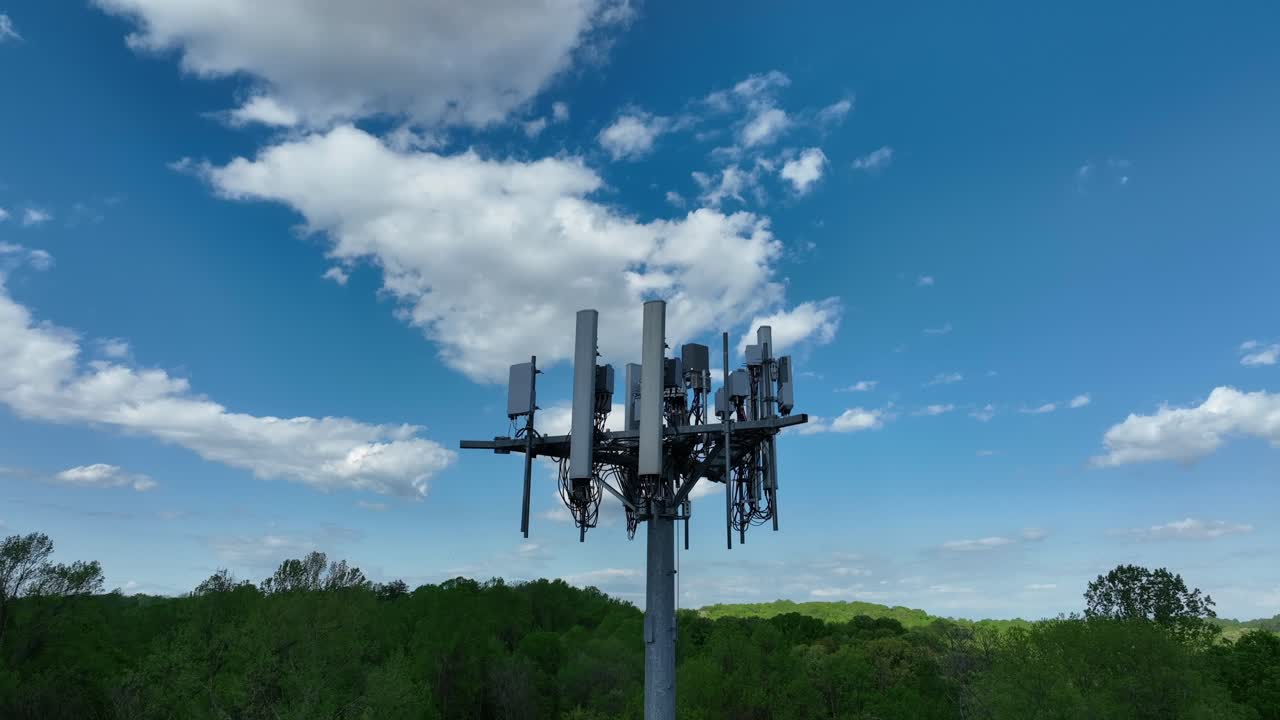 Aerial rising shot of telecommunication tower in rural forest landscape of USA. White clouds on blue sky. 5G cellular wireless connectivity theme.