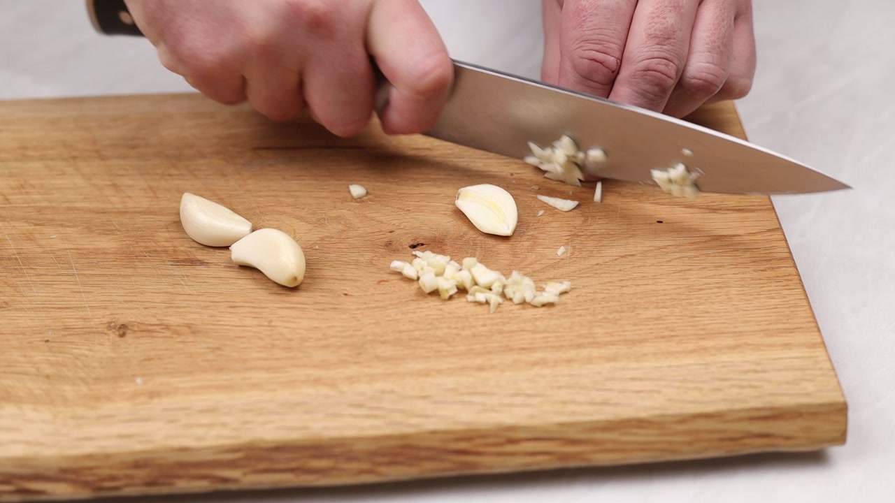 Close up of hand mincing garlic with knife on wooden cutting board during food preparation