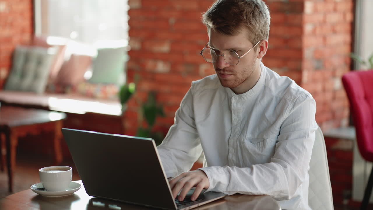 concentrado joven hipster freelancer hombre con barba en gafas sentado en la mesa y mirando la computadora portátil enfocado tipo trabajando desde casa escribiendo mensajes respondiendo correos electrónicos