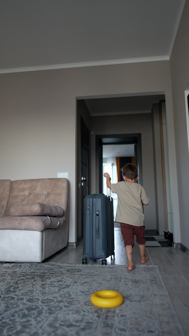 Rear view of a small kid wearing sunglasses and pushing a big grey suitcase. Toddler baby leaves the room with a valise. Vertical video.
